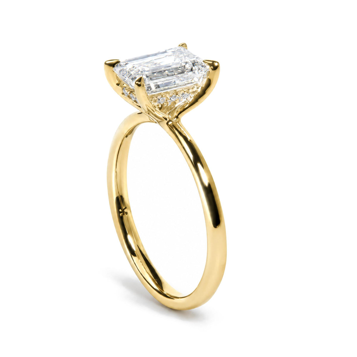 Close-up side angle of a yellow metal ring with a prong-set white emerald-shaped gemstone center surrounded by smaller round white gemstones, shown on a white background.