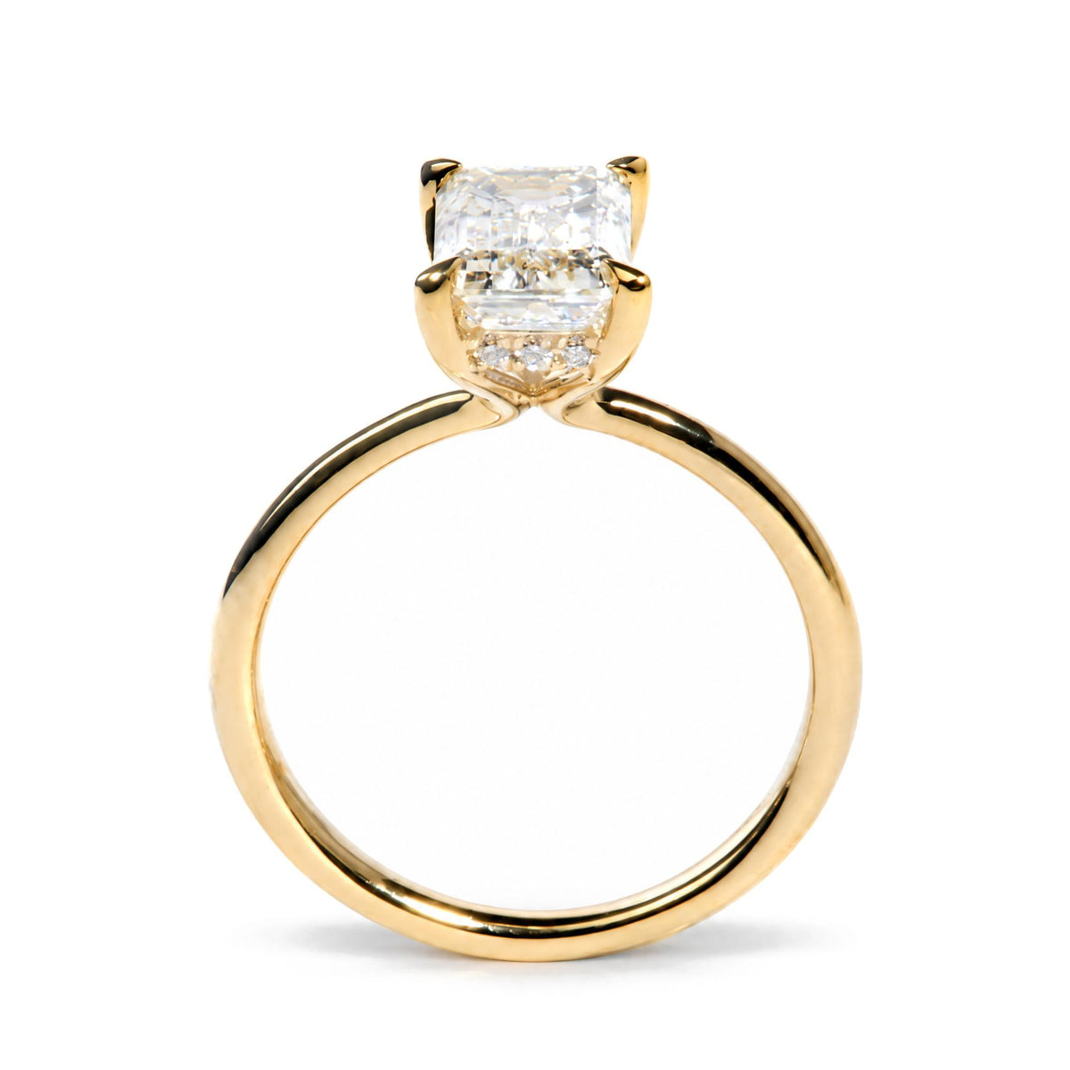 Close-up front angle of a yellow metal ring featuring one emerald-shaped white gemstone in a prong setting, surrounded by multiple small round white gemstones in prong settings.