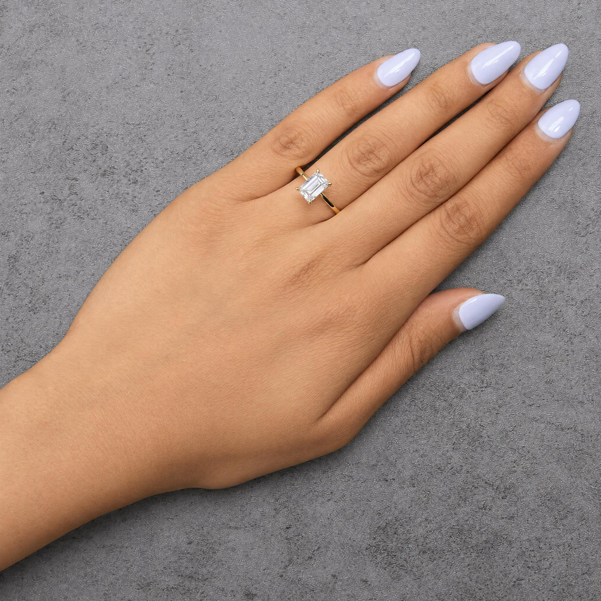 Yellow metal ring with a central emerald-shaped white gemstone and smaller round white gemstones on the band, shown worn on a model's hand from a top-down angle.