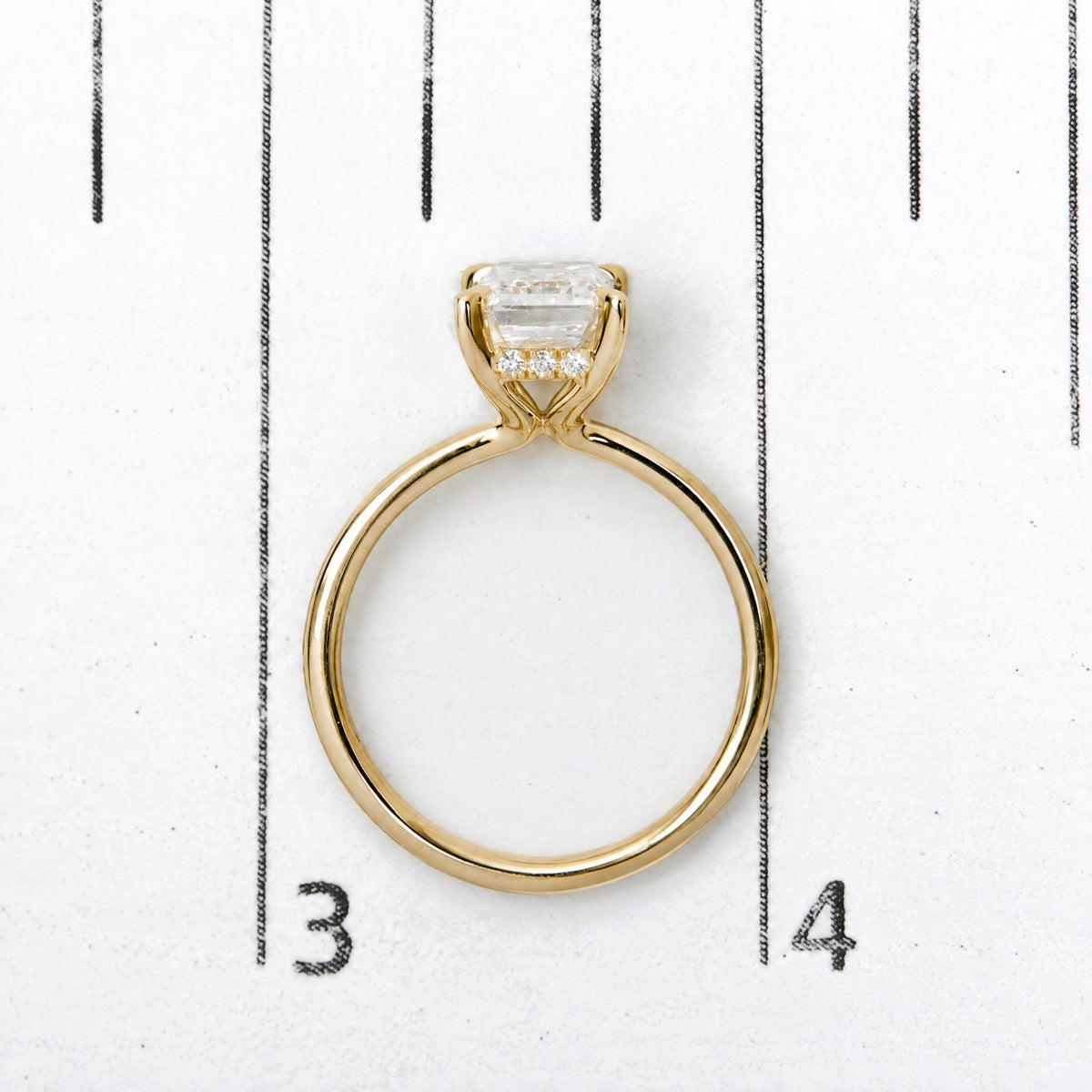 Close-up front view of a yellow metal ring with a central emerald-shaped white gemstone in a prong setting and small round white gemstones beneath it, shown on a numbered scale.