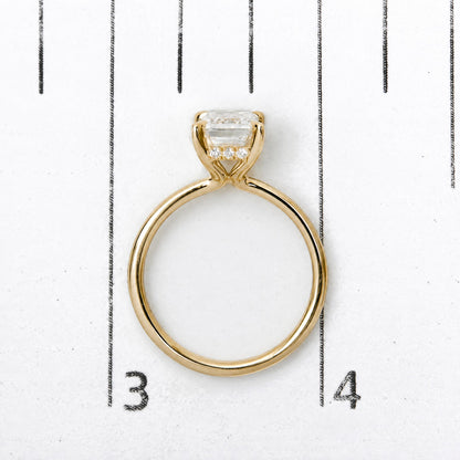 Close-up front view of a yellow metal ring with a central emerald-shaped white gemstone in a prong setting and small round white gemstones beneath it, shown on a numbered scale.