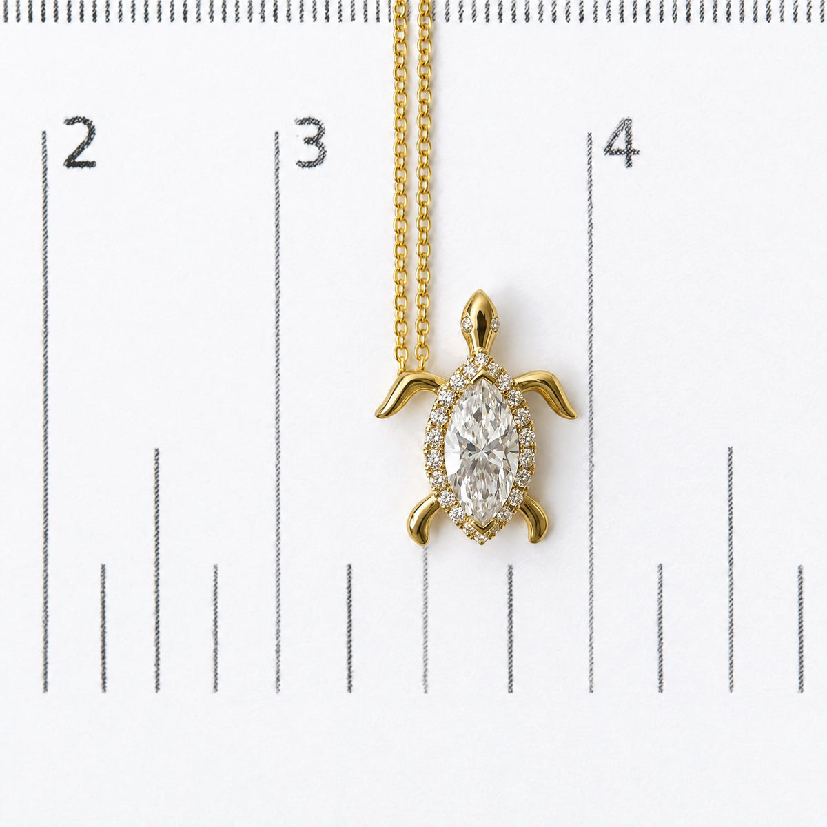 Close-up front view of a yellow metal turtle-shaped necklace pendant with a large marquise-shaped white gemstone center surrounded by round white gemstones, shown on a measurement scale.