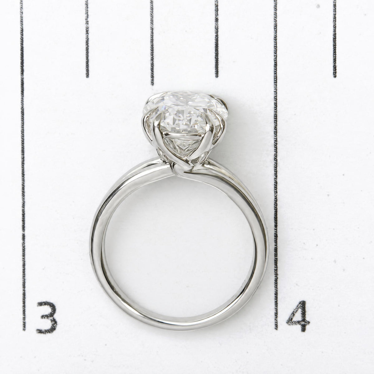 Close-up image of a white ring with an oval-shaped white gemstone in a double claw prong setting, shown on a scale from a top-down angle.