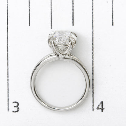 Close-up image of a white ring with an oval-shaped white gemstone in a double claw prong setting, shown on a scale from a top-down angle.