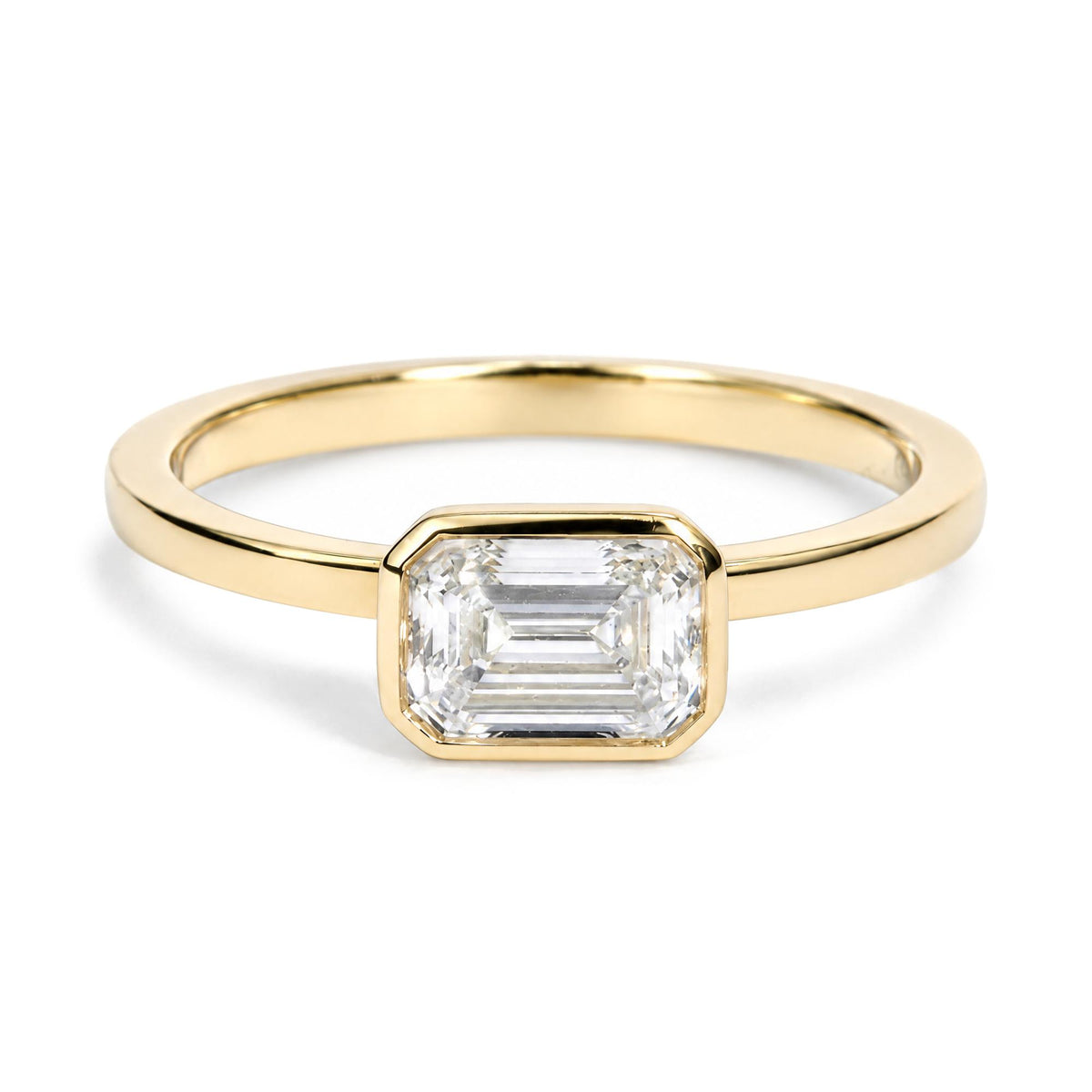 Close-up front view of a yellow metal ring featuring one white emerald-shaped gemstone in a bezel setting.