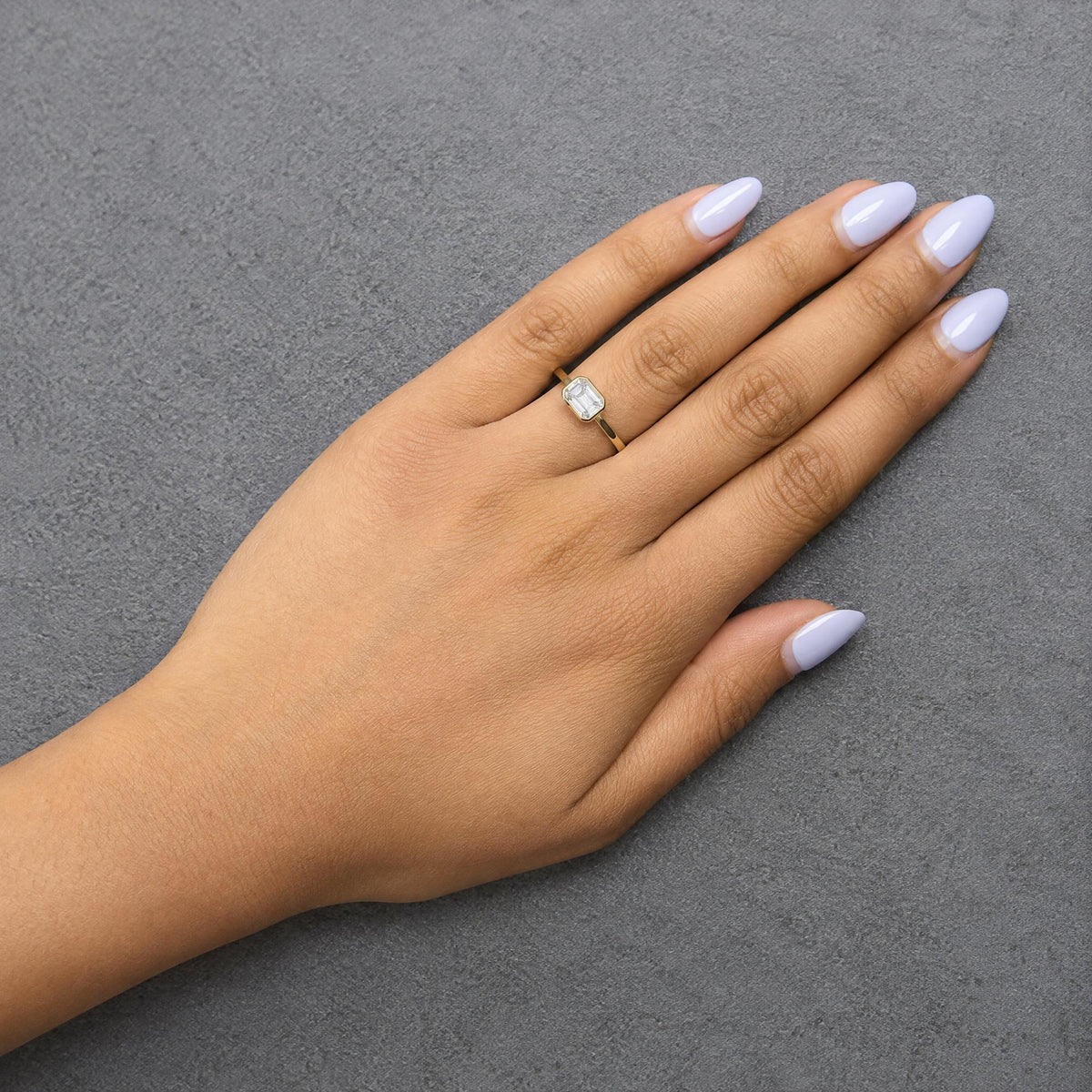 Close-up front angle of a yellow metal ring with a single white emerald-shaped gemstone bezel set, shown worn on a model's finger with light purple nail polish, against a gray textured background.