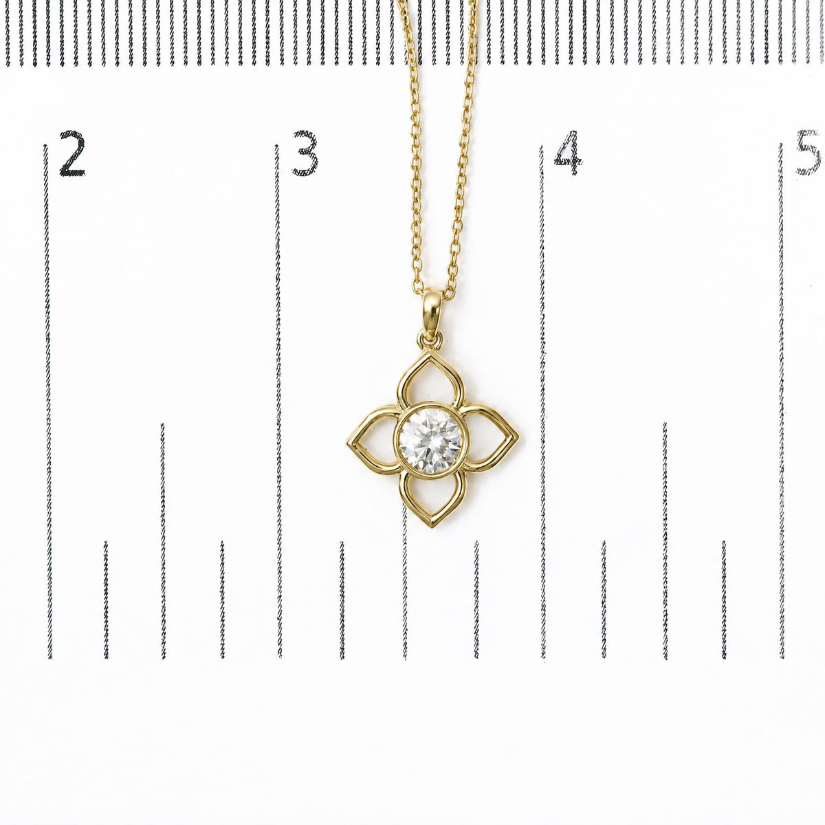 Close-up image of a yellow metal necklace pendant with a clover-shaped design, featuring a round white gemstone in a bezel setting, shown on a measurement scale from a top-down angle.