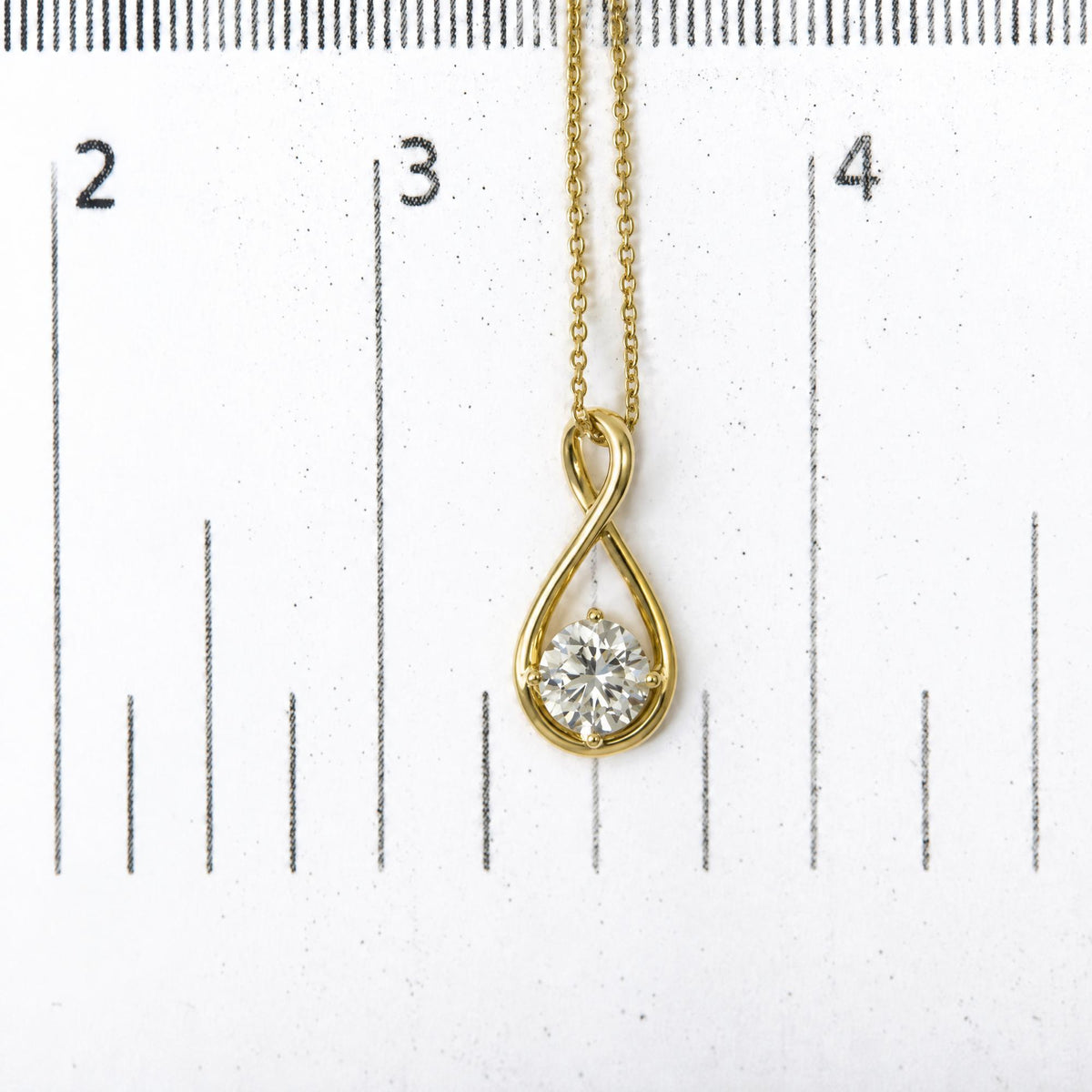 Close-up shot of a yellow metal necklace with an infinity-shaped pendant featuring a round white gemstone in a 4-prong setting, shown on a scale.