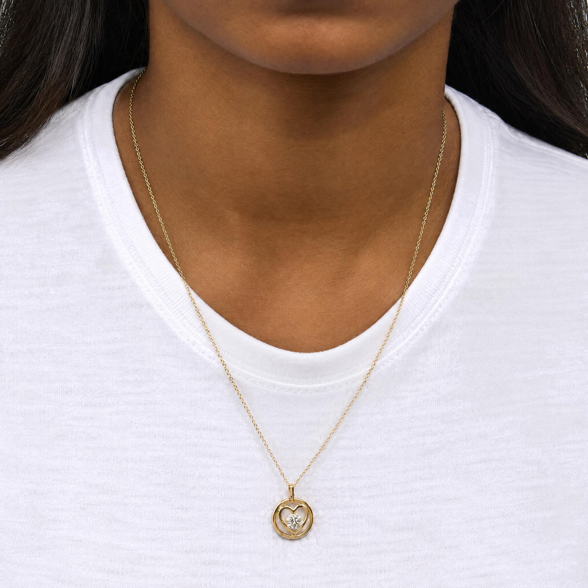 Close-up front view of a model wearing a yellow metal necklace with a round bezel-set white gemstone pendant in a heart-shaped design.