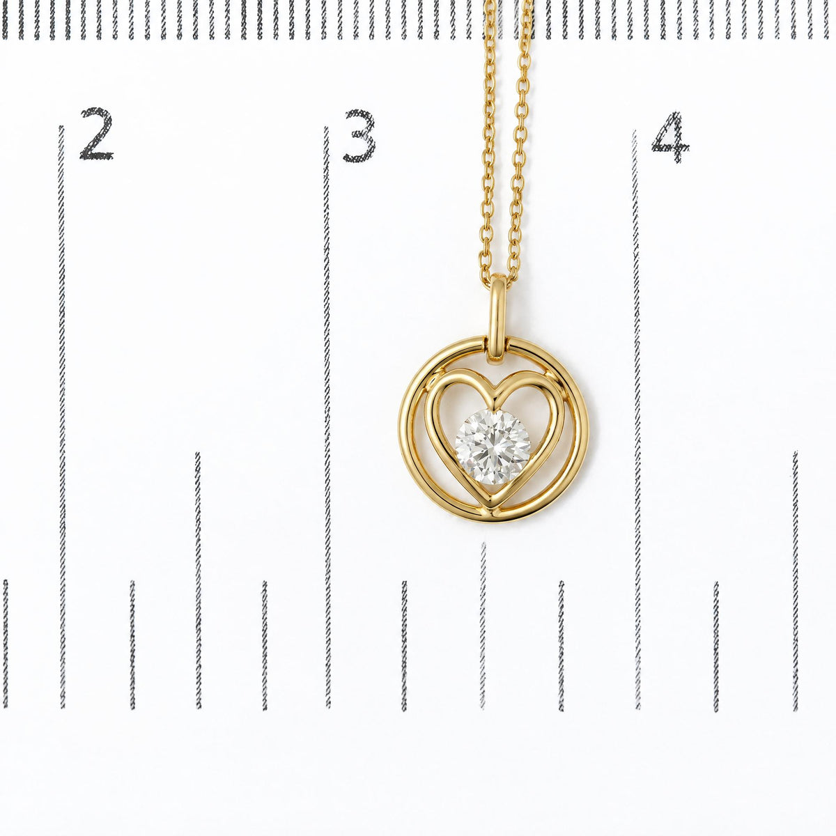 Close-up image of a yellow metal necklace pendant featuring interlocking circular and heart shapes with a round white gemstone in a bezel setting at the center, shown on a measurement scale.