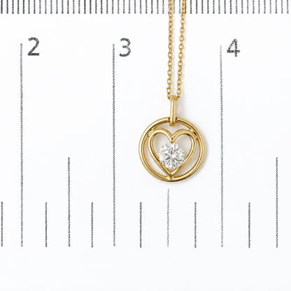 Close-up image of a yellow metal necklace pendant featuring interlocking circular and heart shapes with a round white gemstone in a bezel setting at the center, shown on a measurement scale.