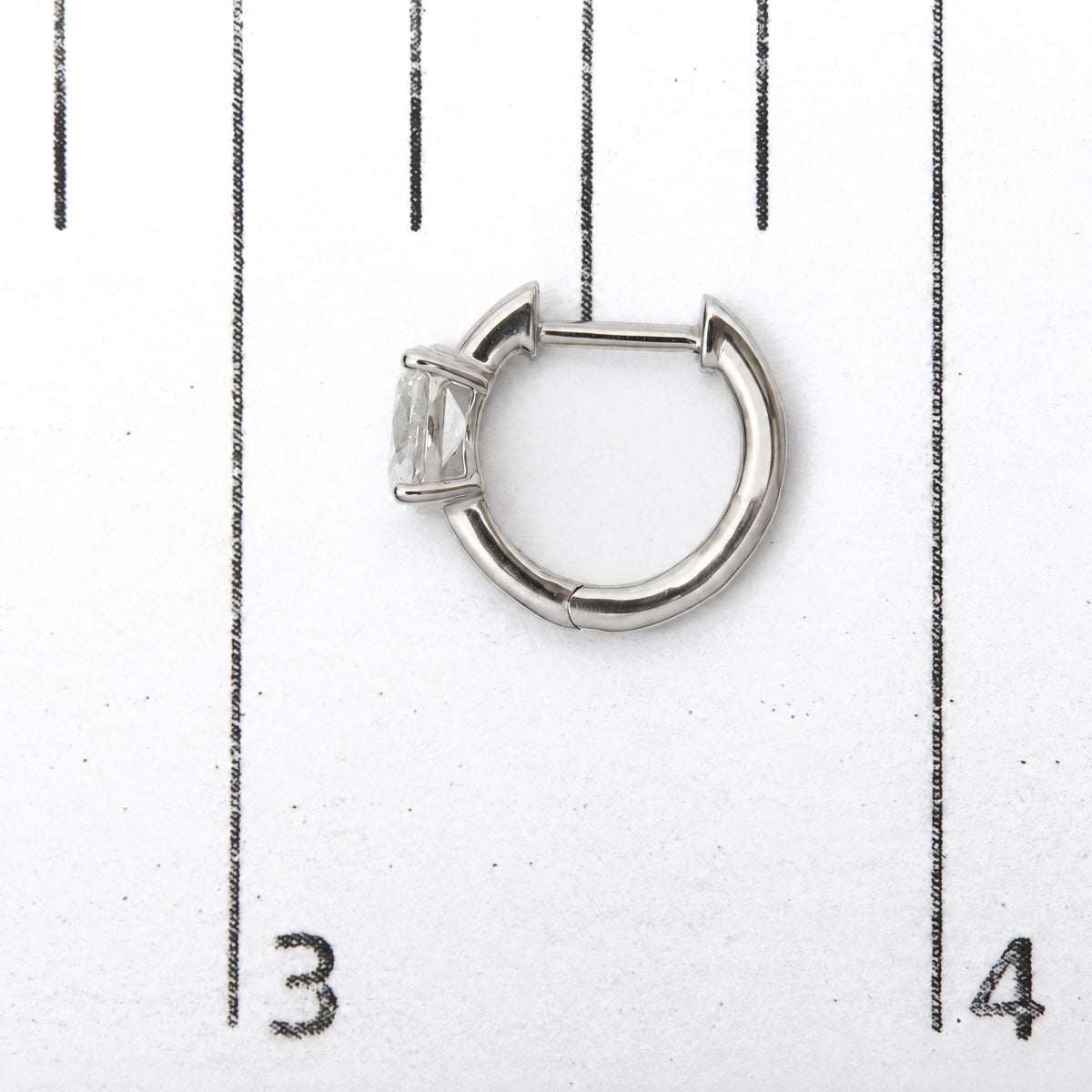 Close-up top view of a white metal hoop earring with two oval white gemstones in a four-prong setting, shown on a scale.
