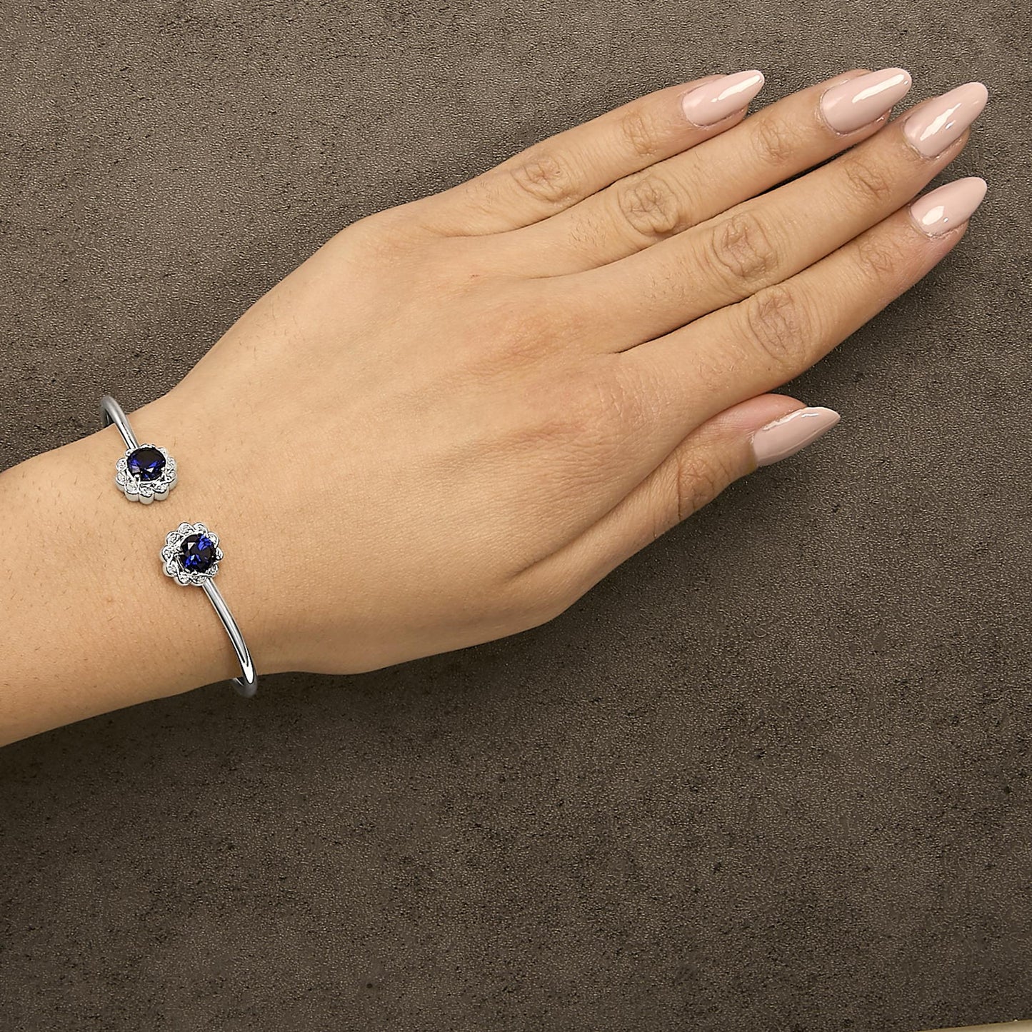 White metal open cuff bracelet with two round blue gemstones surrounded by round white gemstones in prong settings, shown worn on a model's wrist, angled top-down.