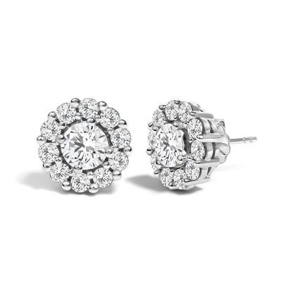 Pair of white metal stud earrings with a central round white gemstone in four-prong setting, surrounded by smaller round white gemstones in shared prong setting, shown from a front and side angle on a white background.
