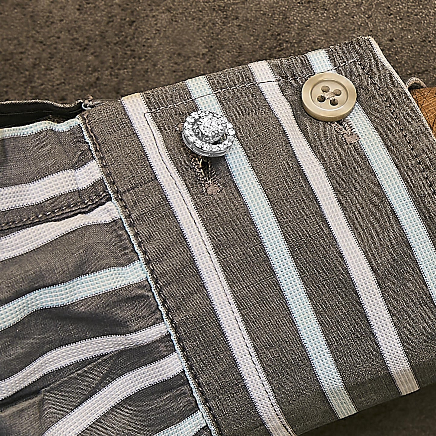 Close-up angled view of a white metal cuff link adorned with round white gemstones in a 4-prong setting, worn on a striped fabric cuff with a beige button.
