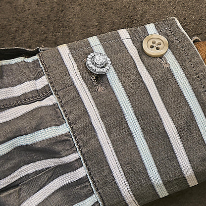 Close-up angled view of a white metal cuff link adorned with round white gemstones in a 4-prong setting, worn on a striped fabric cuff with a beige button.