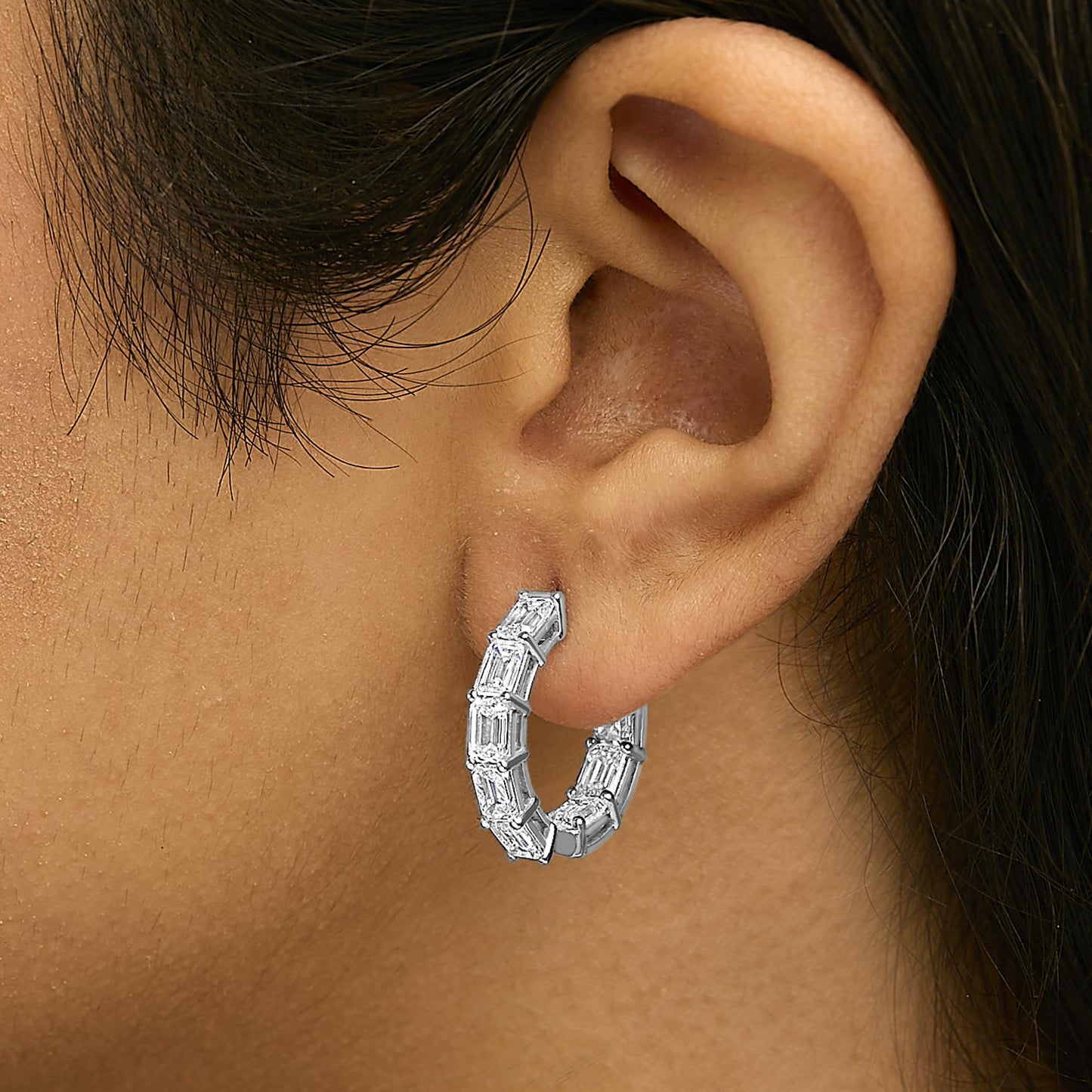 Close-up side view of a model wearing a white metal hoop earring featuring multiple emerald-shaped white gemstones in a shared prong setting.