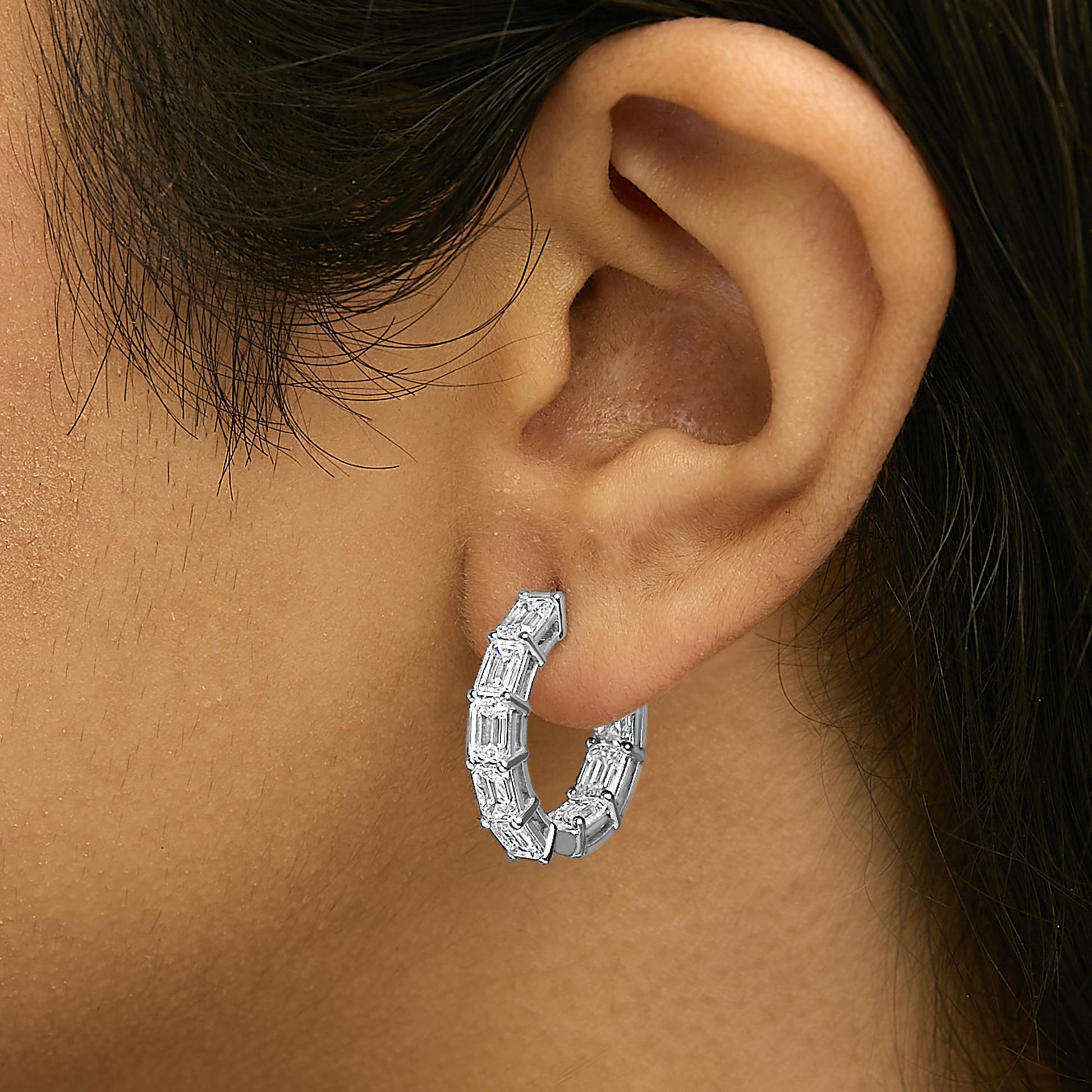 Close-up side view of a model wearing a white metal hoop earring featuring multiple emerald-shaped white gemstones in a shared prong setting.