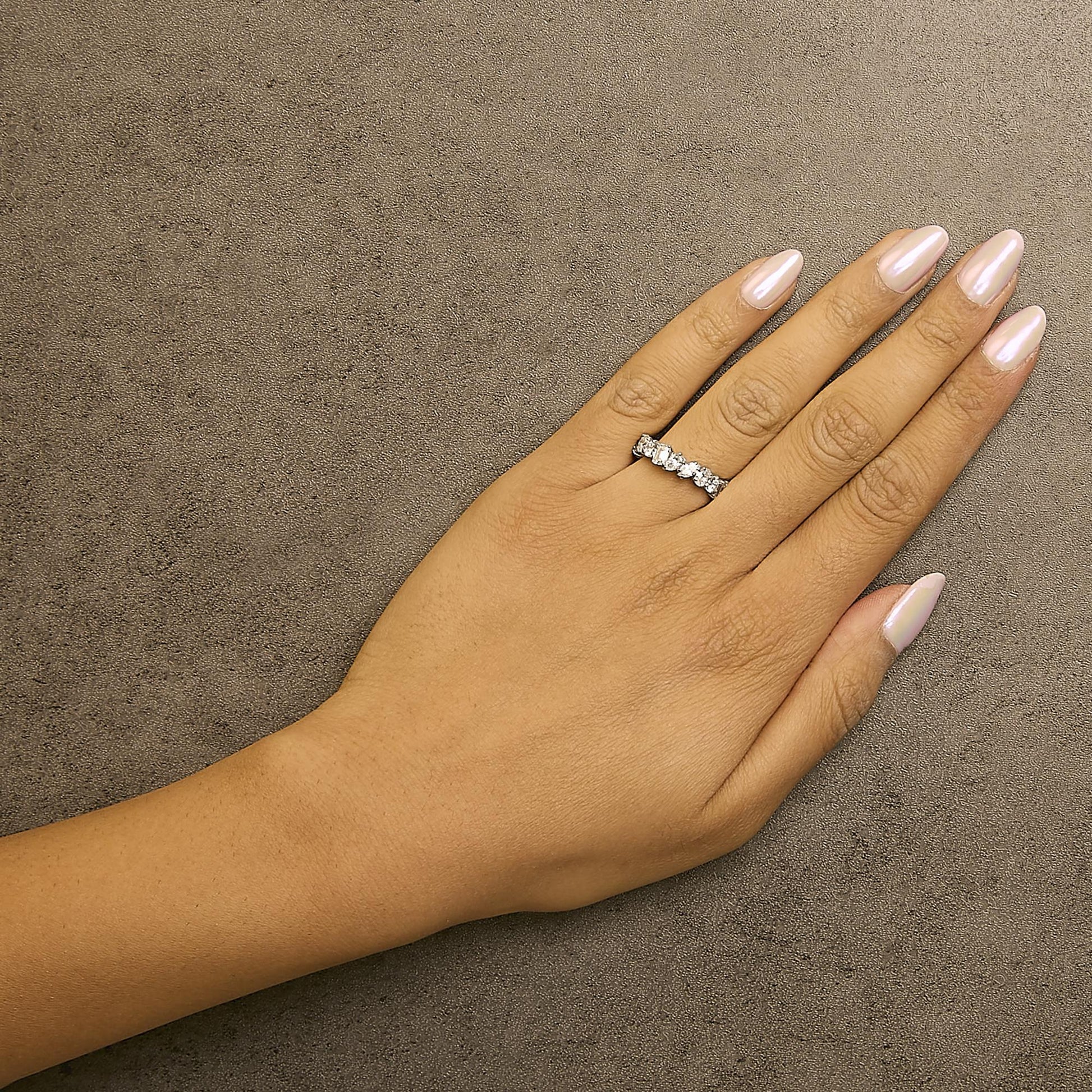 White metal ring with alternating white round and emerald-shaped gemstones in prong settings, shown worn on a model's hand from a top angle.