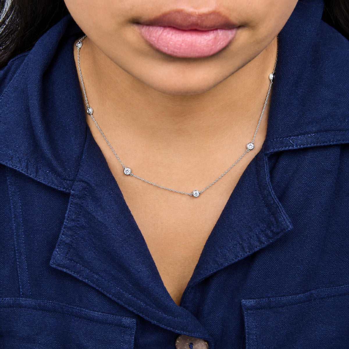 Close-up front view of a model wearing a white metal necklace with round bezel-set white gemstones evenly spaced along the chain.