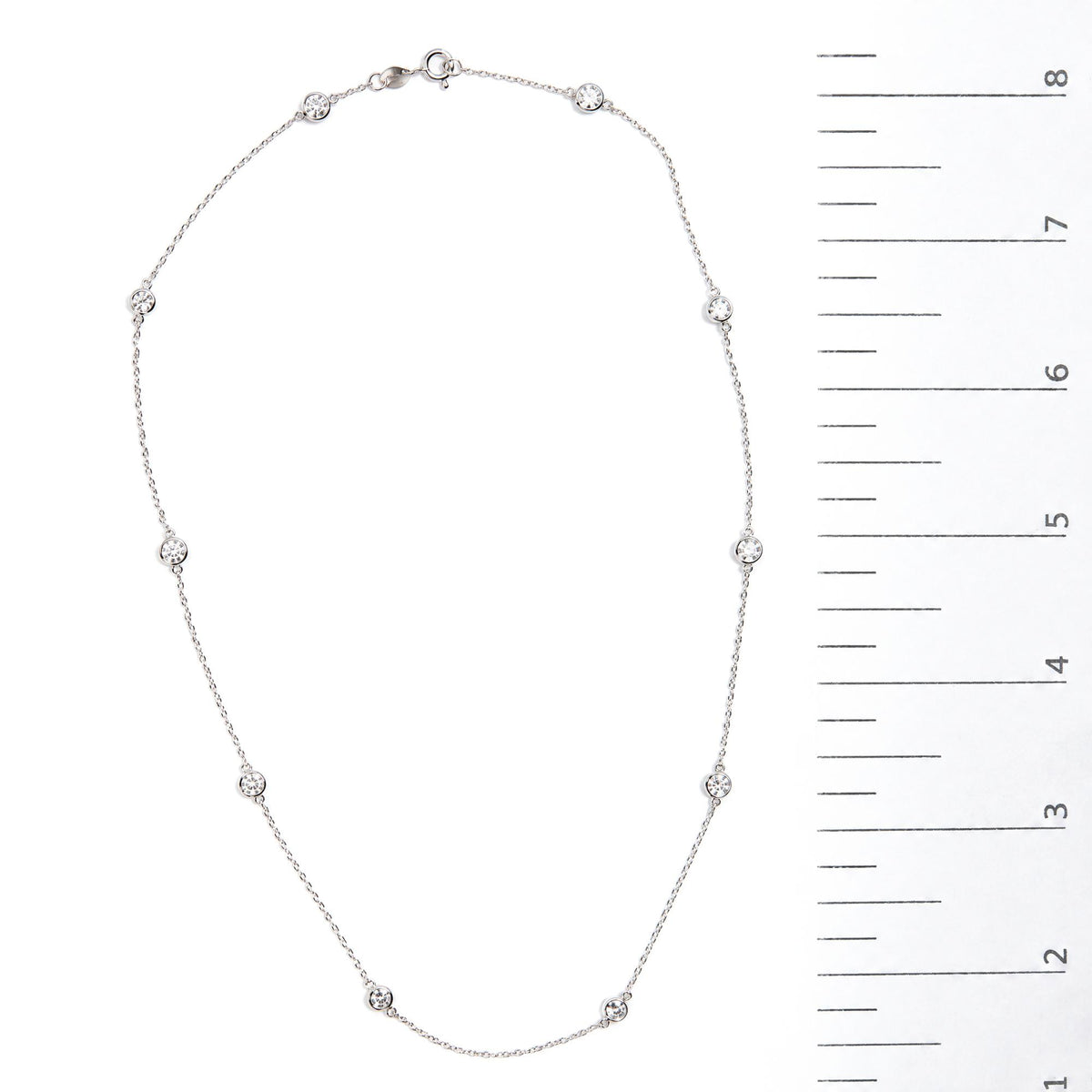 Close-up image of a white necklace with round bezel-set white gemstones evenly spaced along a thin chain, shown flat on a white surface next to a measurement scale.