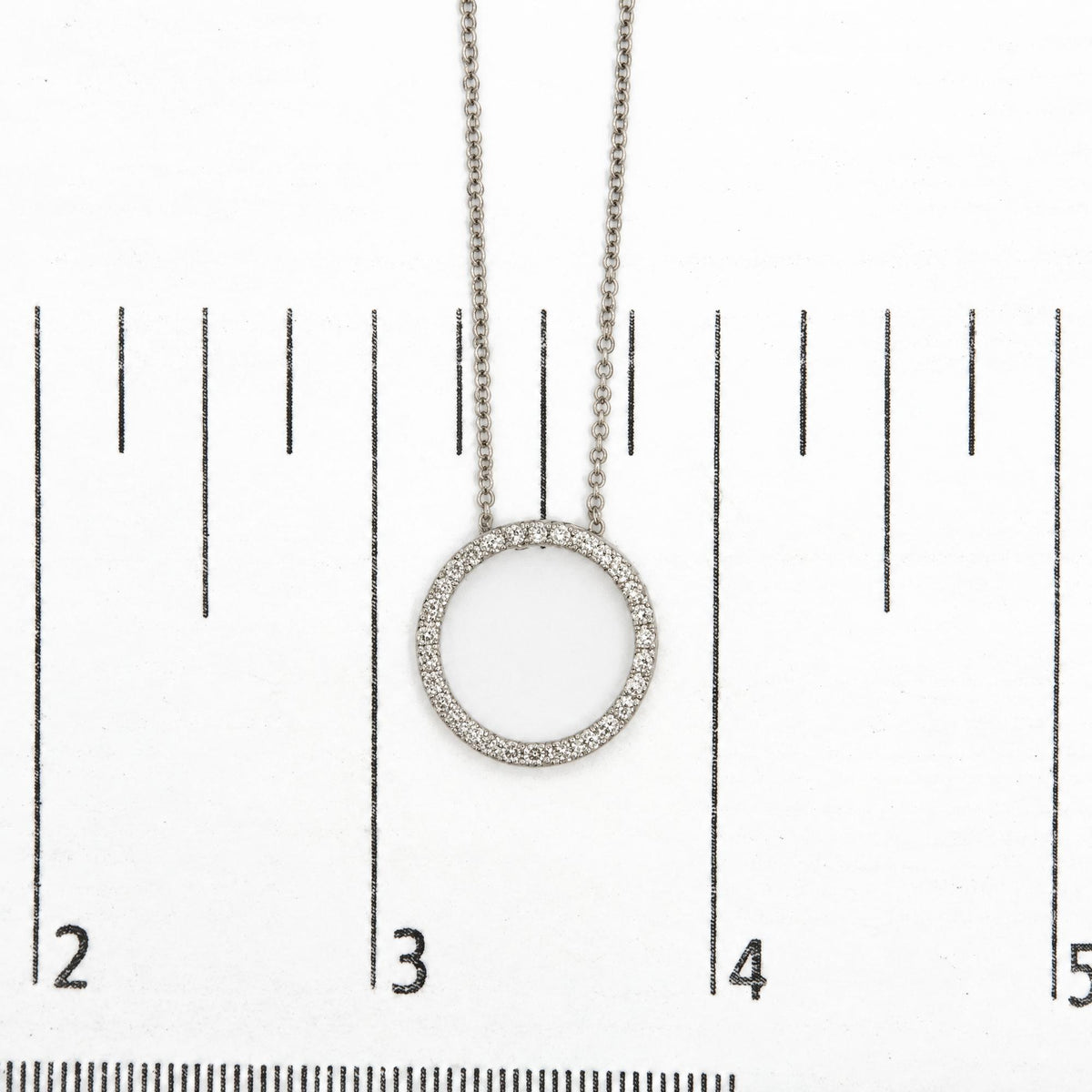 Close-up shot of a white metal circular necklace pendant adorned with round white gemstones in a prong setting, shown on a scale.