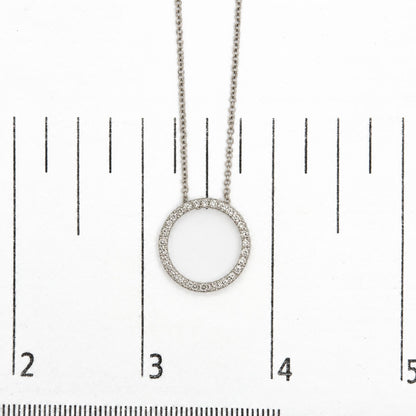 Close-up shot of a white metal circular necklace pendant adorned with round white gemstones in a prong setting, shown on a scale.