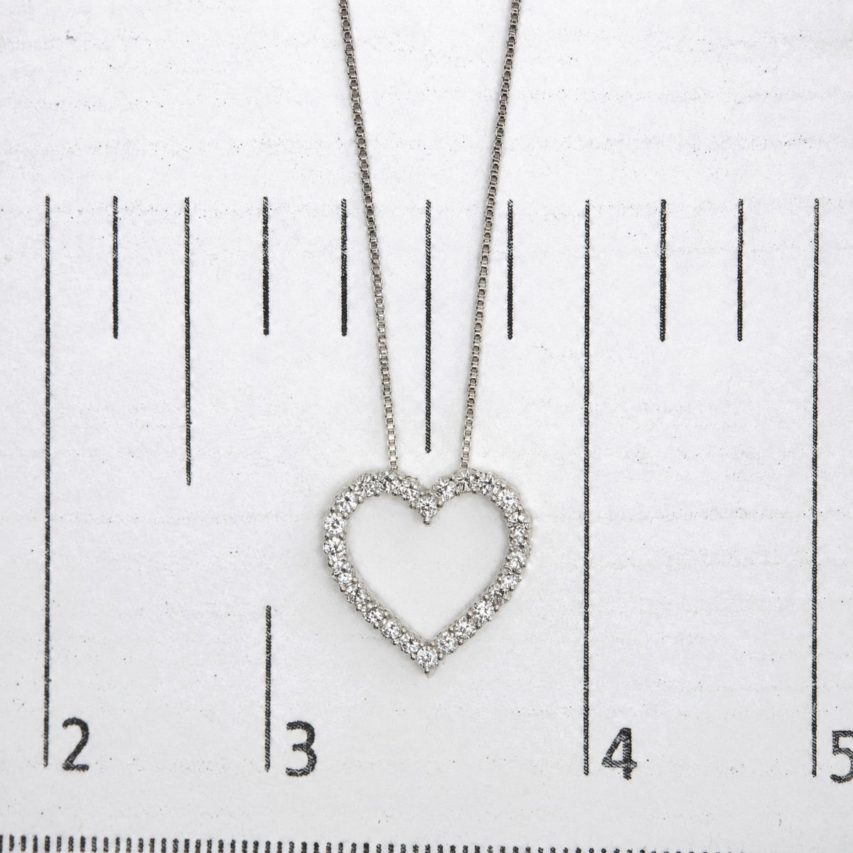 Close-up front view of a white metal heart-shaped necklace pendant with round white gemstones in prong settings, shown on a measurement scale.