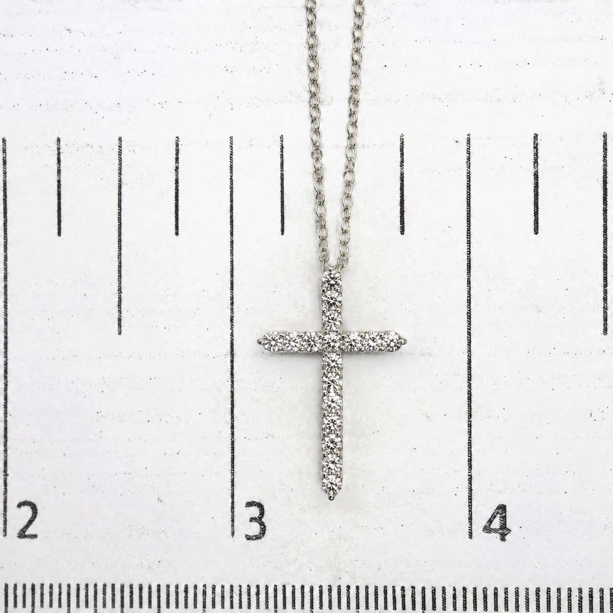 Close-up image of a white metal necklace with a cross pendant covered in white round gemstones in prong settings, shown on a scale.