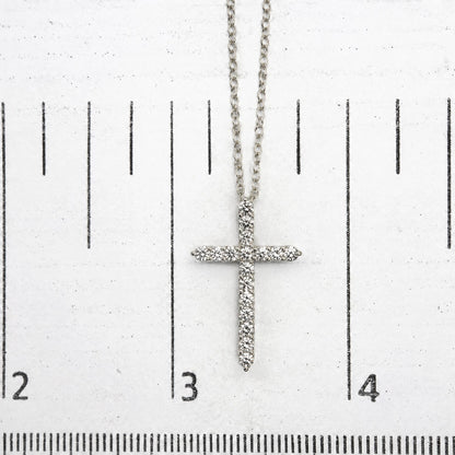 Close-up image of a white metal necklace with a cross pendant covered in white round gemstones in prong settings, shown on a scale.