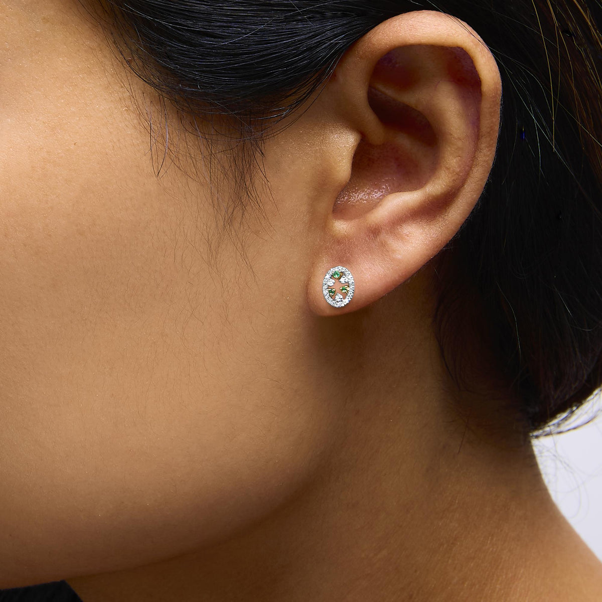 Close-up side view of a model wearing yellow metal oval-shaped earrings featuring prong-set white round gemstones surrounding prong-set green round gemstones.