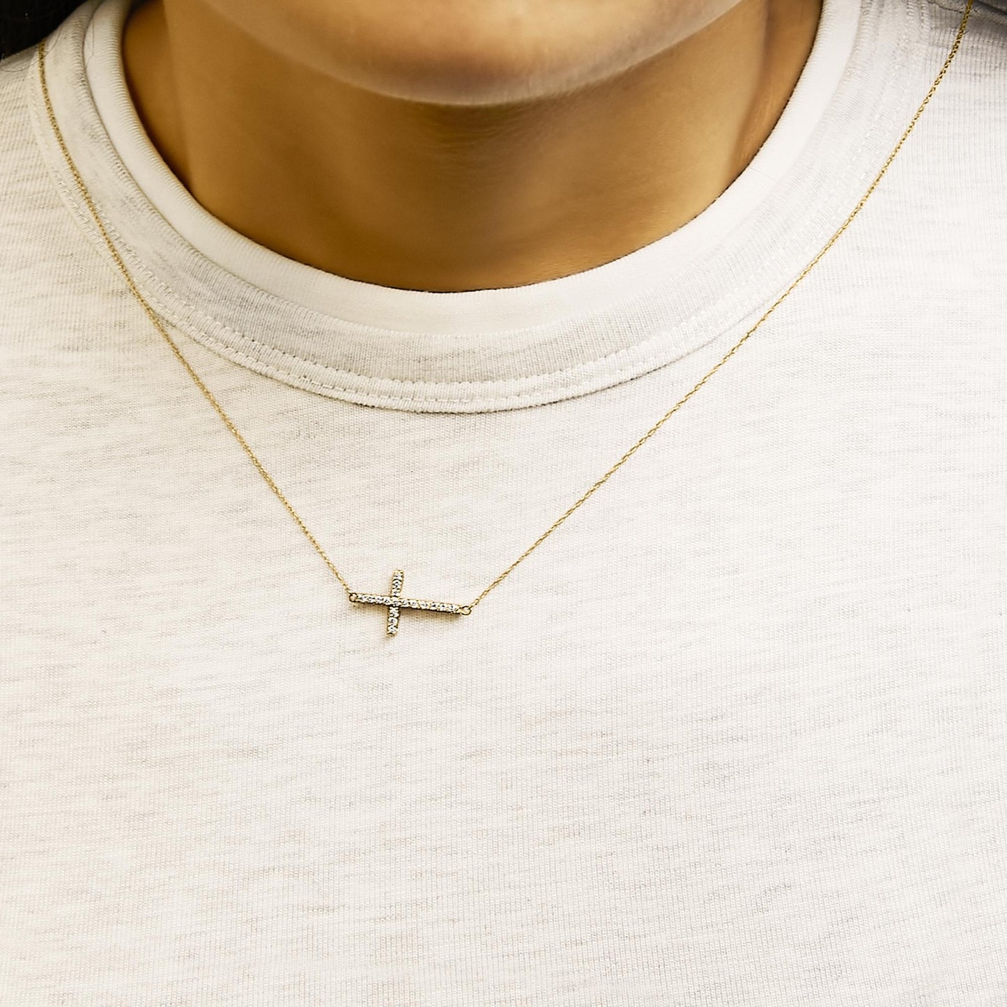 Necklace worn on a model, featuring a horizontal cross pendant with white round gemstones in a shared prong setting on a delicate yellow chain, shown from a front close-up angle.