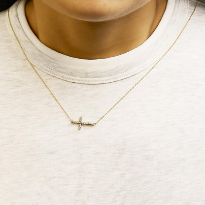 Necklace worn on a model, featuring a horizontal cross pendant with white round gemstones in a shared prong setting on a delicate yellow chain, shown from a front close-up angle.