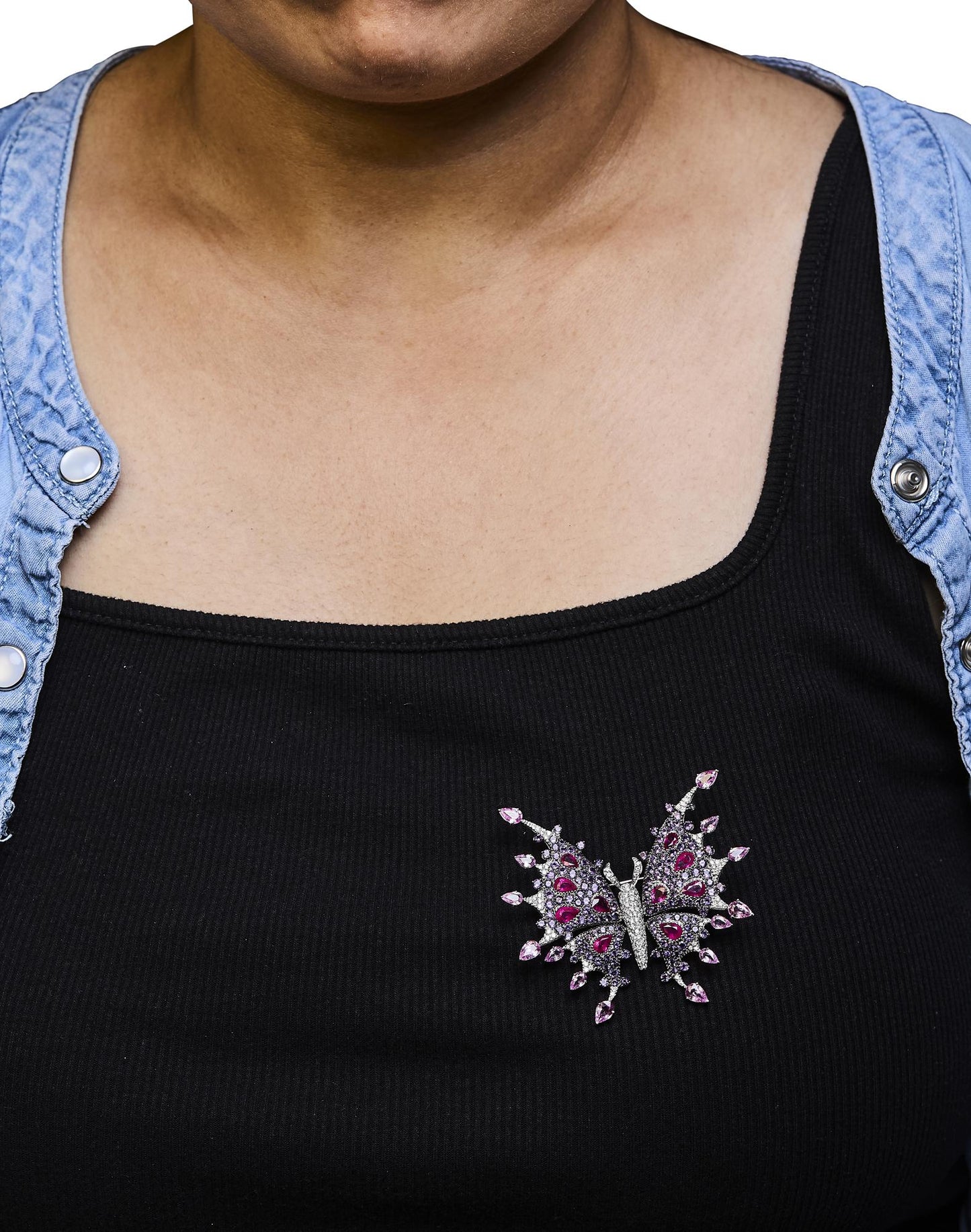 Brooch shaped like a butterfly with white metal, white gemstones, red pear-shaped gemstones, and pink pear-shaped gemstones, pinned on black top worn by a model, front-facing angle.