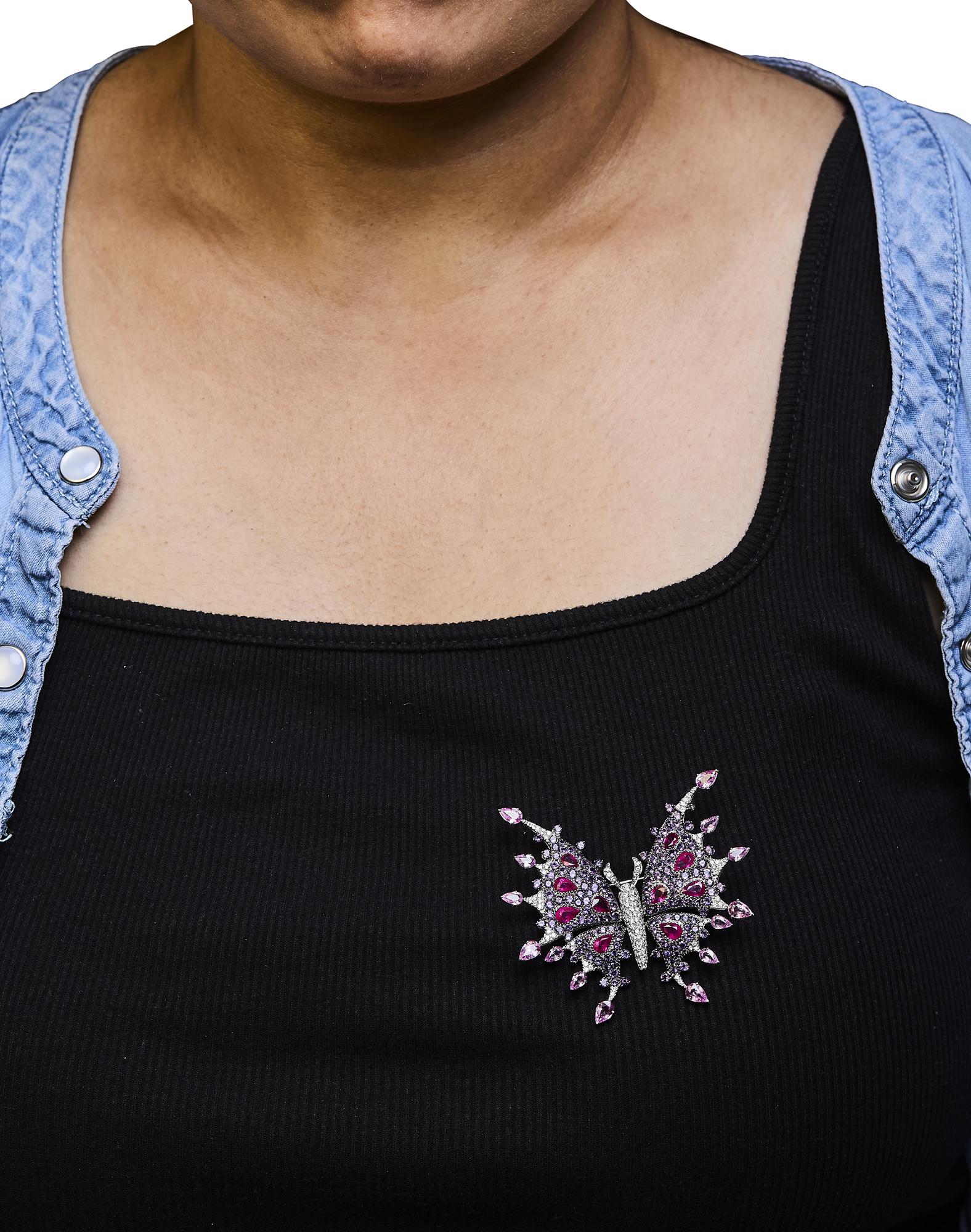 Brooch shaped like a butterfly with white metal, white gemstones, red pear-shaped gemstones, and pink pear-shaped gemstones, pinned on black top worn by a model, front-facing angle.