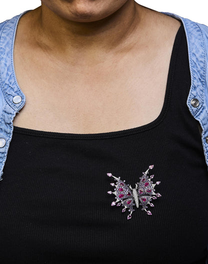 Brooch shaped like a butterfly with white metal, white gemstones, red pear-shaped gemstones, and pink pear-shaped gemstones, pinned on black top worn by a model, front-facing angle.