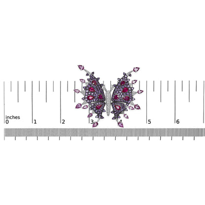 Top-down view of a white metal butterfly-shaped brooch with white round pave-set gemstones on the body, purple round pave-set gemstones and red and pink pear-shaped gemstones on the wings, placed on a ruler for size reference.