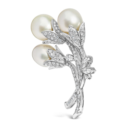 White metal brooch shaped like a flower bouquet with three large white pearls and multiple small round white gemstones in a pave setting, shown in a side angle view.