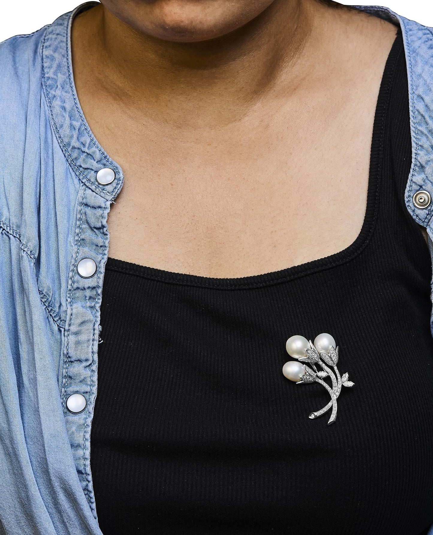 White metal brooch with three pearl-like gemstones and white round pave-set gemstones arranged as leaves and stems, shown pinned on black top worn by a model, front angle.