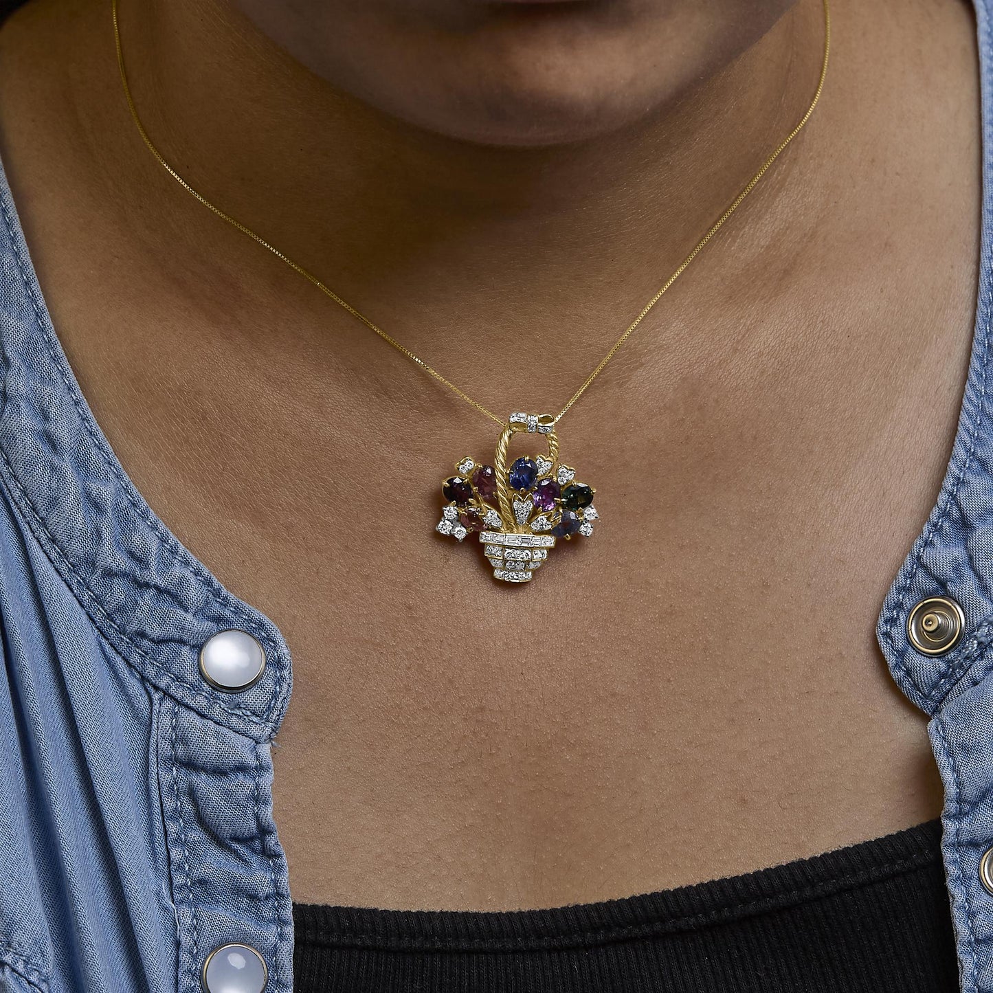 Yellow metal brooch shaped like a basket with decorative floral design, featuring round and baguette-shaped white gemstones, a blue oval gemstone, and a purple pear-shaped gemstone, worn on a model, shown from a front angle.
