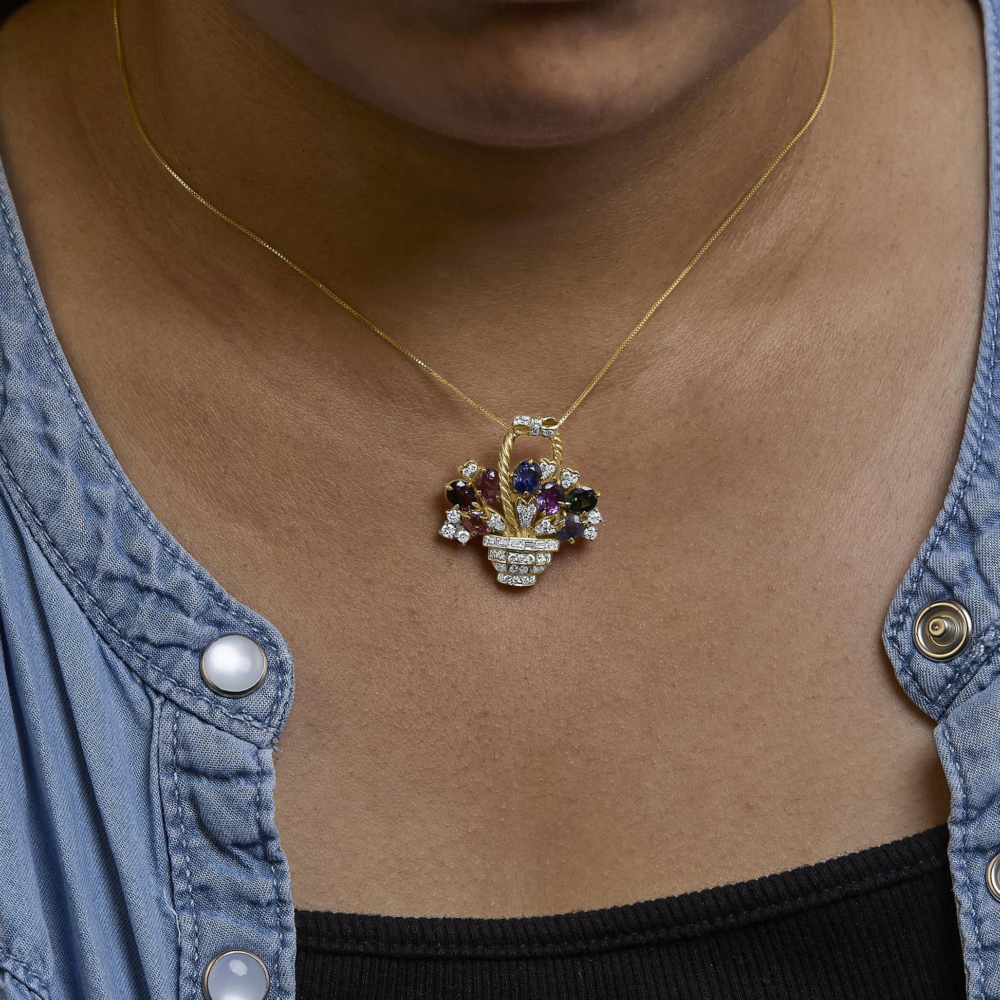 Yellow metal brooch shaped like a basket with decorative floral design, featuring round and baguette-shaped white gemstones, a blue oval gemstone, and a purple pear-shaped gemstone, worn on a model, shown from a front angle.