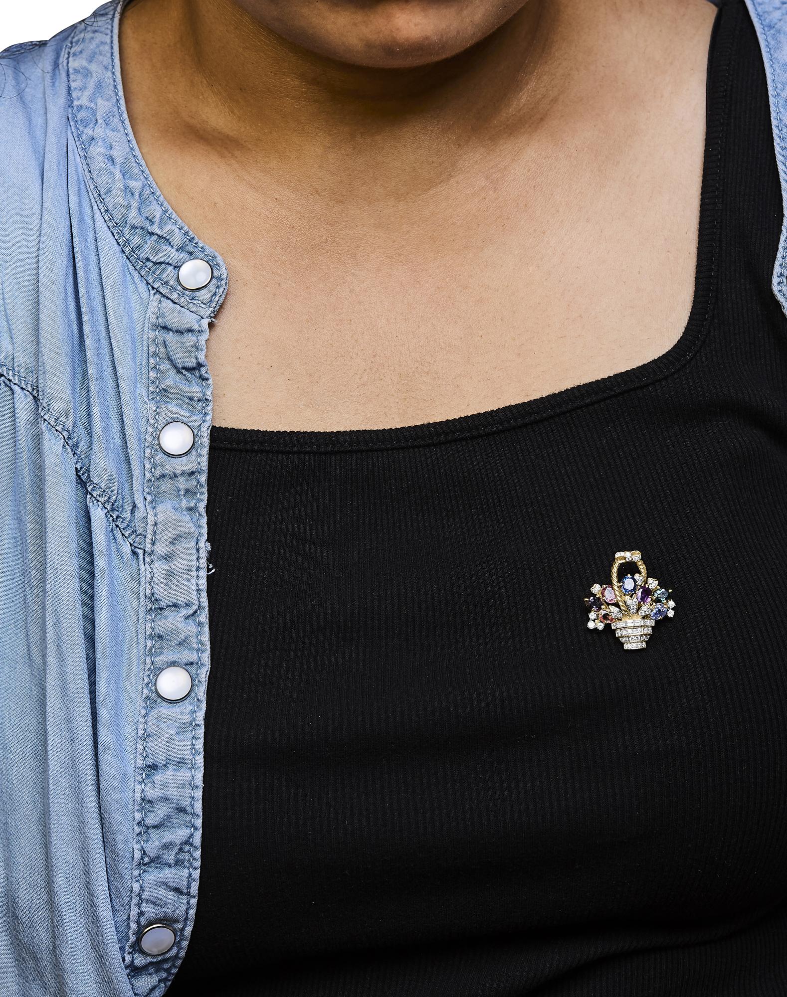 Yellow metal brooch with blue oval gemstone, purple pear-shaped gemstone, white round and baguette-shaped gemstones, worn on chest of a model, front view.