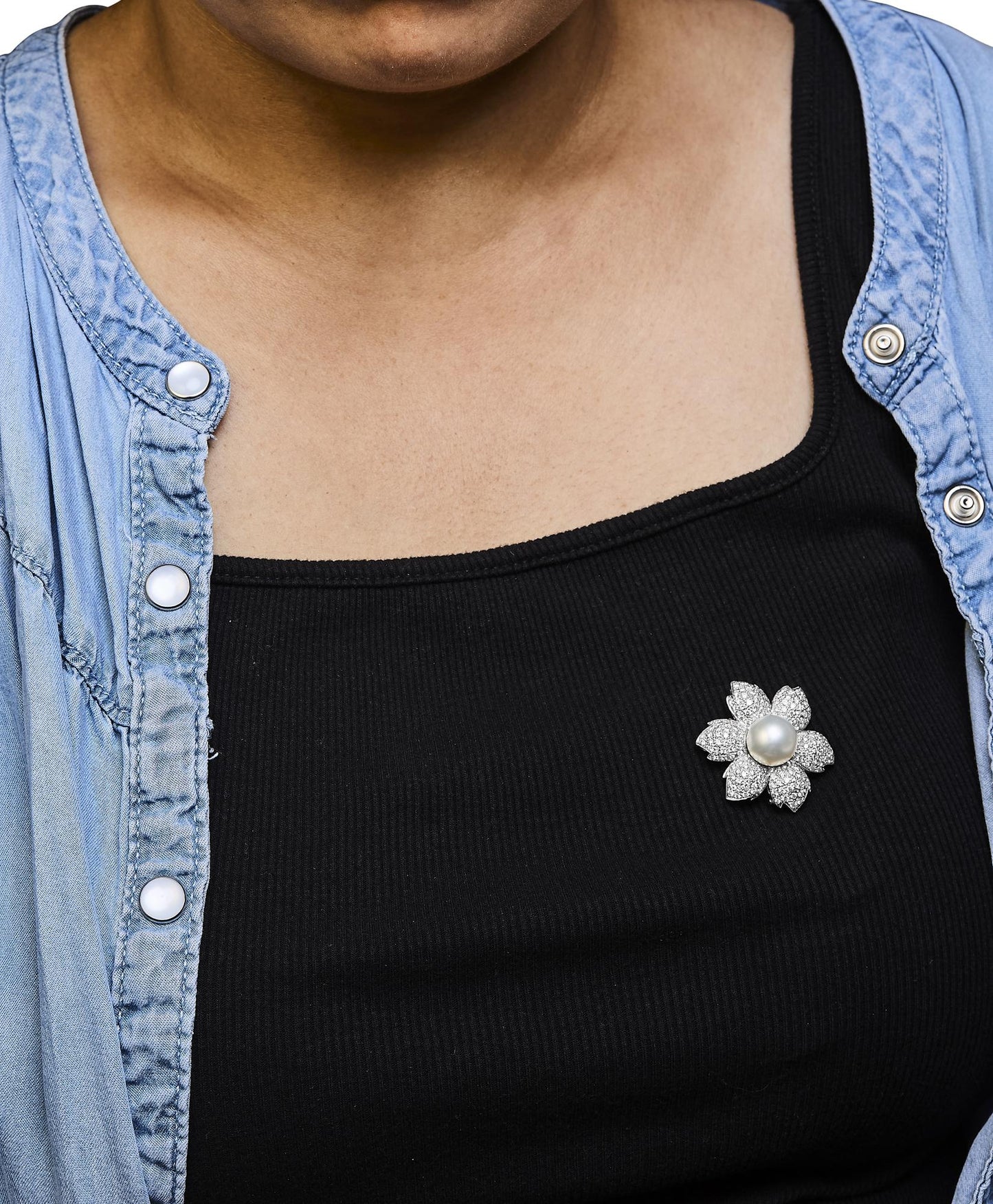 Close-up front view of a white metal flower-shaped brooch with petals paved with round white gemstones and a central white gemstone, worn on a black top by a model.