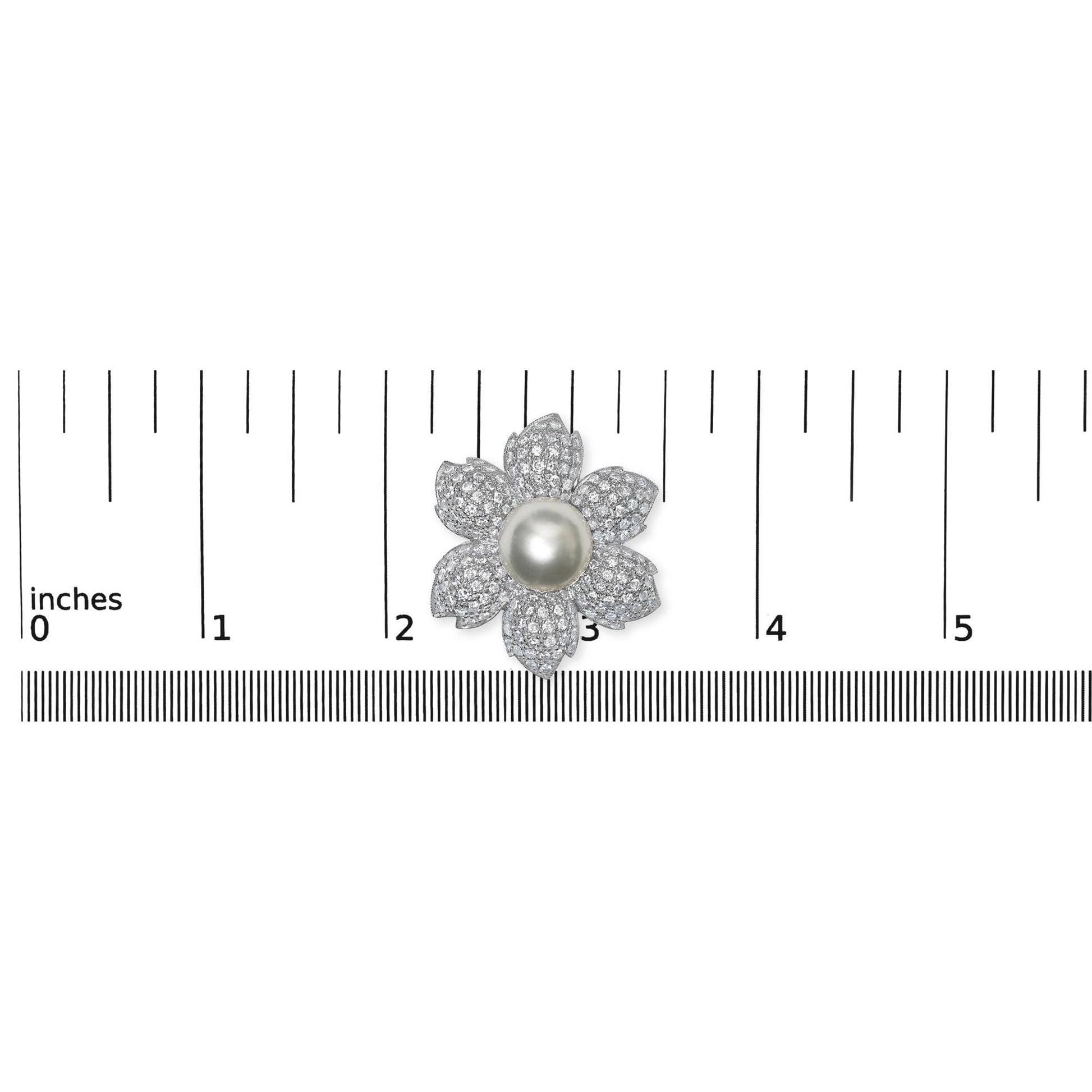 Top-down view of a white metal flower-shaped brooch with a central smooth round white gemstone and petals encrusted with small round white gemstones in pave setting, placed on a ruler for size reference.