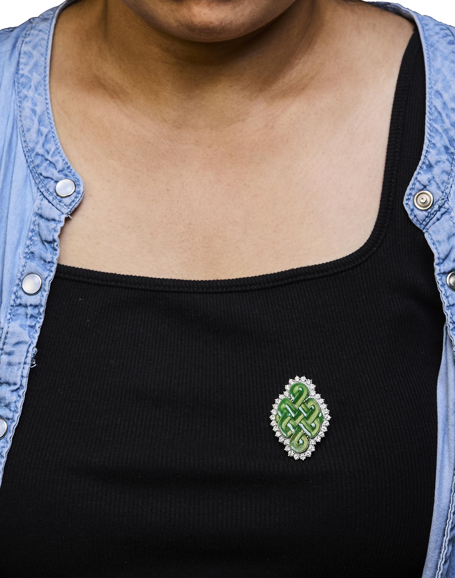 White metal brooch with a green gemstone center in a fantasy shape, surrounded by round white gemstones in prong settings, worn on a model, front angle.