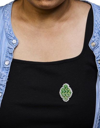 White metal brooch with a green gemstone center in a fantasy shape, surrounded by round white gemstones in prong settings, worn on a model, front angle.