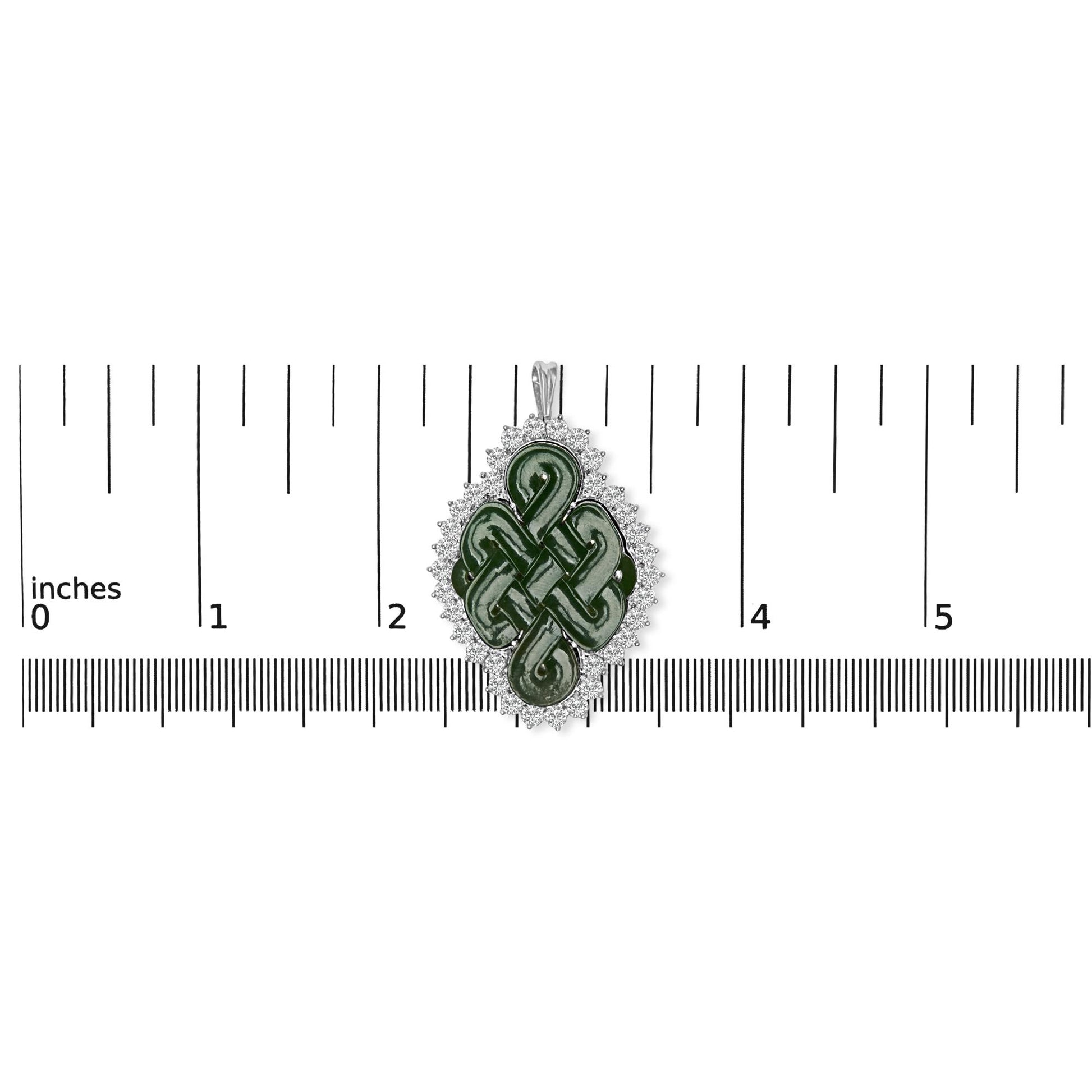 Top-down view of a white metal brooch with a green fantasy-shaped gemstone in the center, surrounded by a border of round white gemstones, placed on a ruler for size reference.
