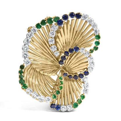 Yellow metal brooch with a swirling leaf design, accented with round white gemstones, blue gemstones, and green gemstones, shown at a slight front angle.
