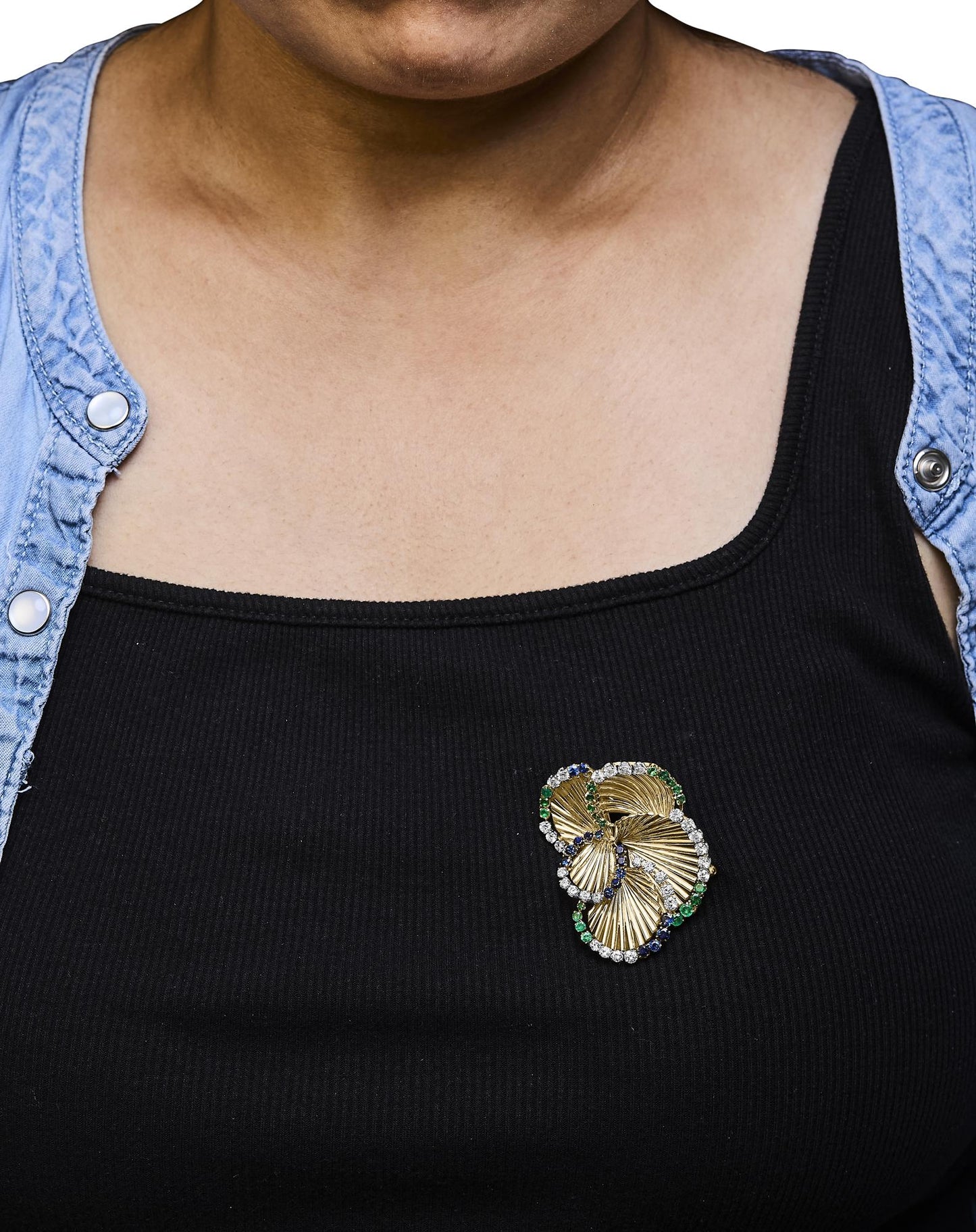 Yellow metal brooch with white, blue, and green round gemstones in 4-prong settings, worn on a model's black top, shot from front angle.