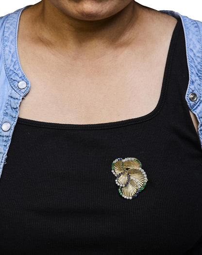 Yellow metal brooch with white, blue, and green round gemstones in 4-prong settings, worn on a model's black top, shot from front angle.